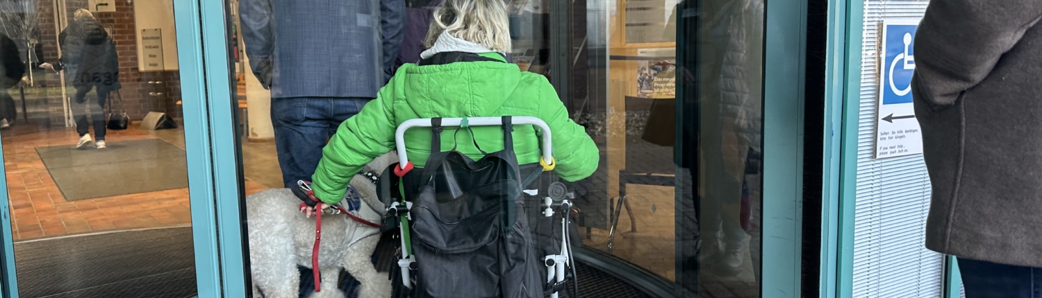 Person im Rollstuhl mit Hund betritt ein Gebäude durch eine automatische Drehtür.