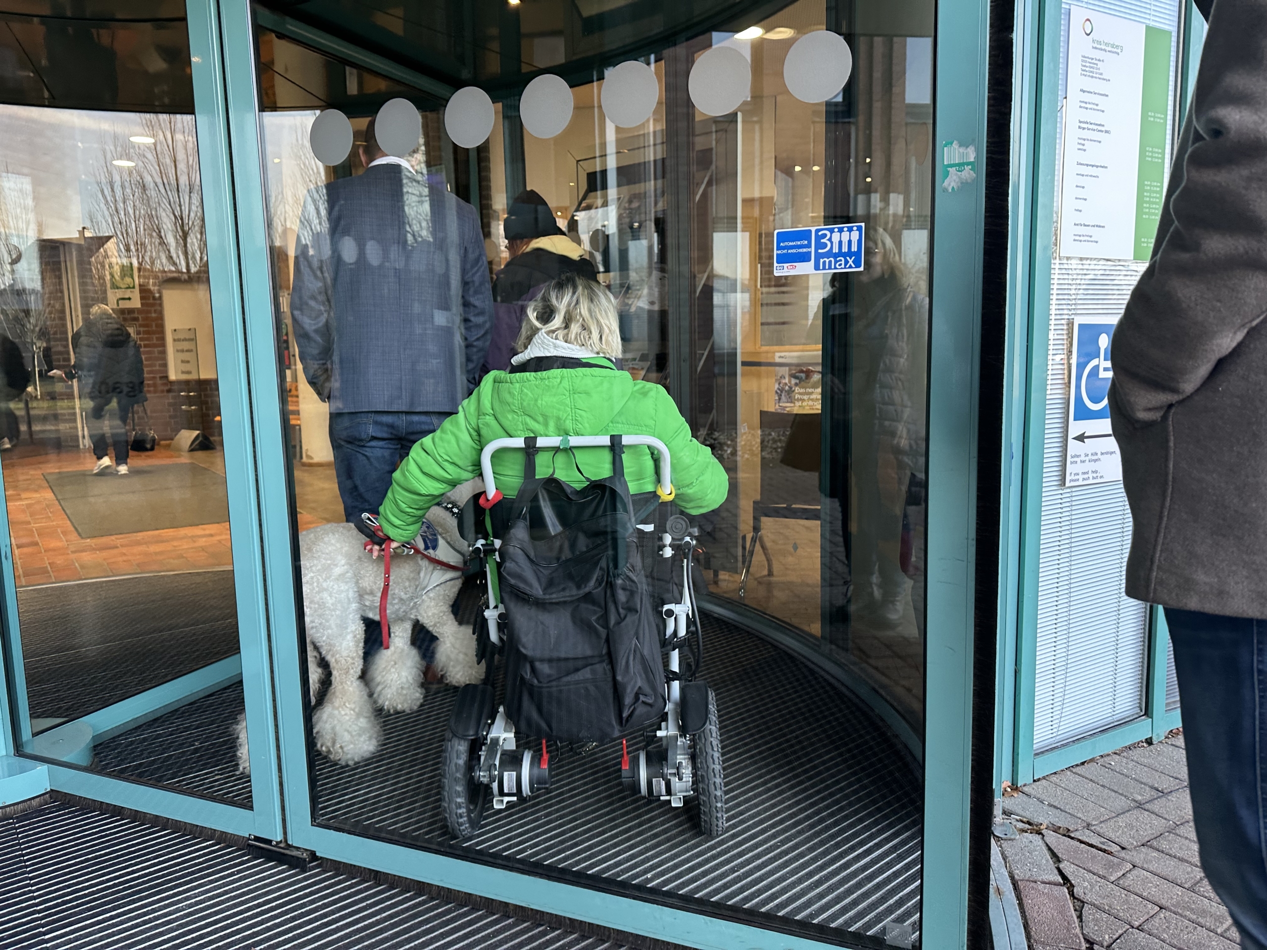 Person im Rollstuhl mit Hund betritt ein Gebäude durch eine automatische Drehtür.