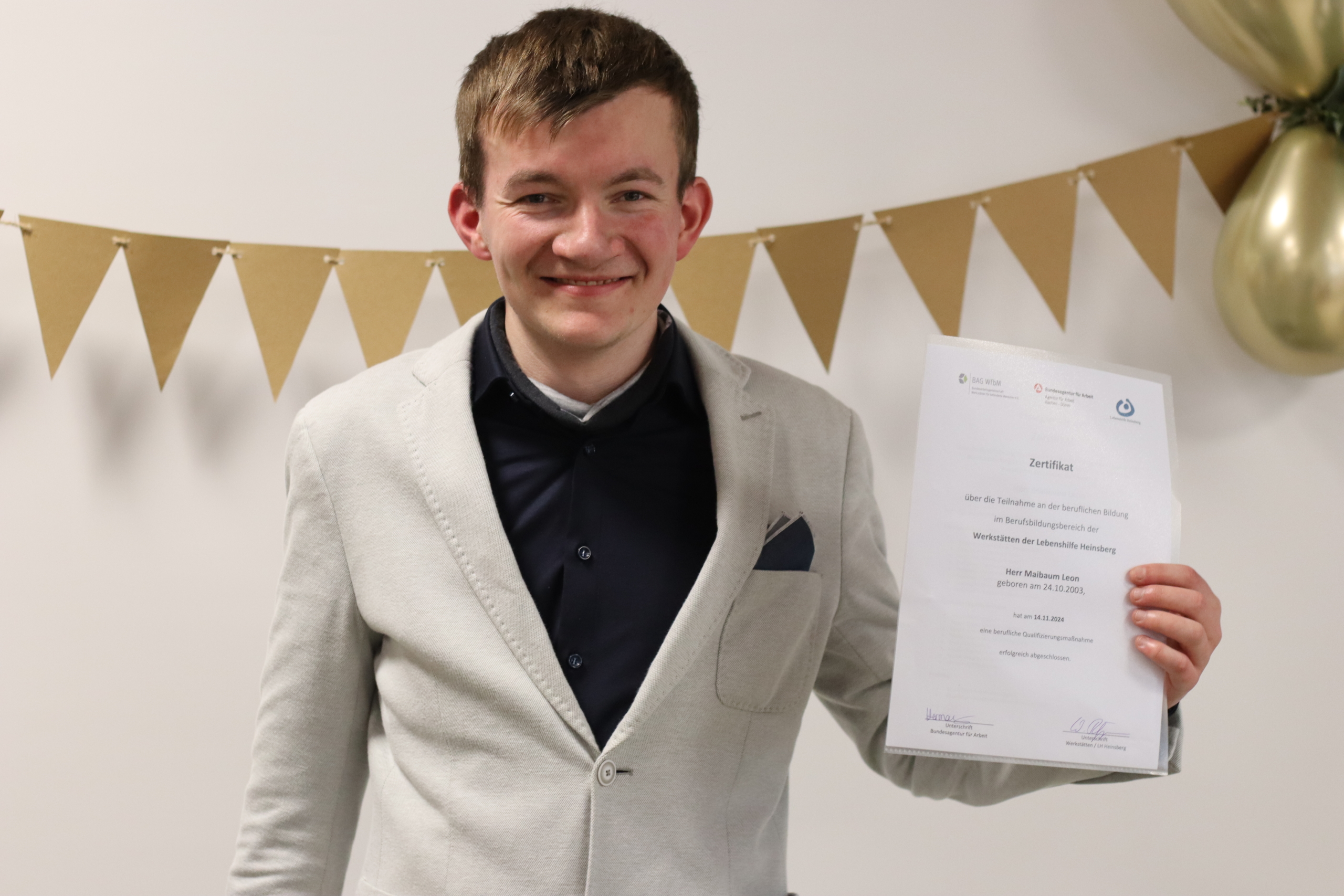 Ein Mann hält ein Dokument. Er trägt einen Anzug und lächelt. Hinter ihm sind bunte Girlanden.