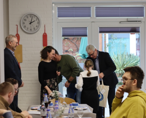Menschen sitzen an Tischen mit Essen. Zwei Männer sprechen miteinander. Eine Frau steht daneben.