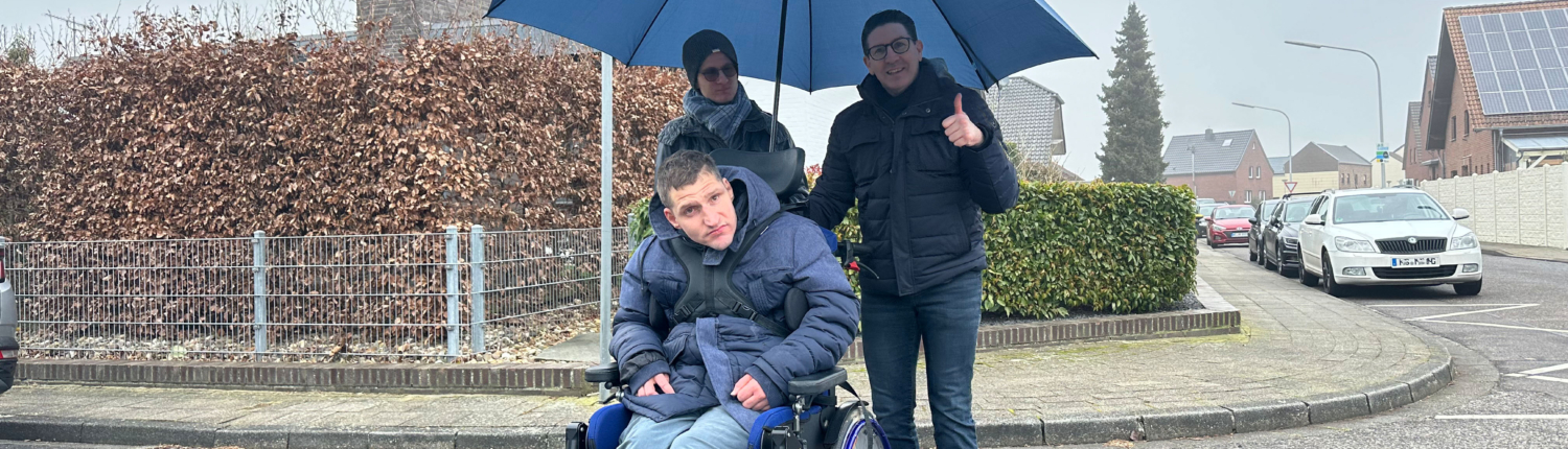 Ein Mann sitzt im Rollstuhl und geht mit einem Freund. Es regnet, deshalb halten sie einen Schirm.