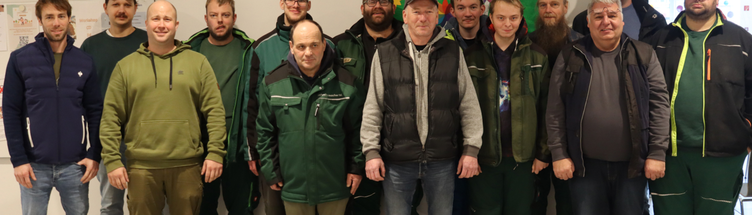 Gruppenfoto von Männern in Arbeitskleidung, die zusammenstehen. Im Hintergrund ist eine grüne Wand mit bunten Elementen zu sehen. Die Personen scheinen aus dem Bereich der Landwirtschaft oder Forstwirtschaft zu stammen.