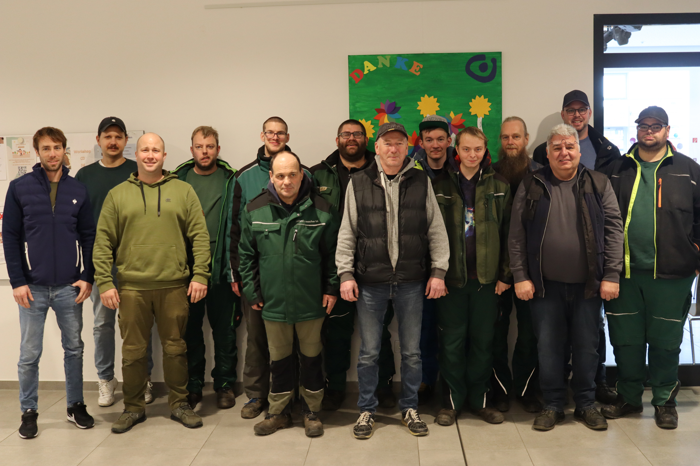 Gruppenfoto von Männern in Arbeitskleidung, die zusammenstehen. Im Hintergrund ist eine grüne Wand mit bunten Elementen zu sehen. Die Personen scheinen aus dem Bereich der Landwirtschaft oder Forstwirtschaft zu stammen.