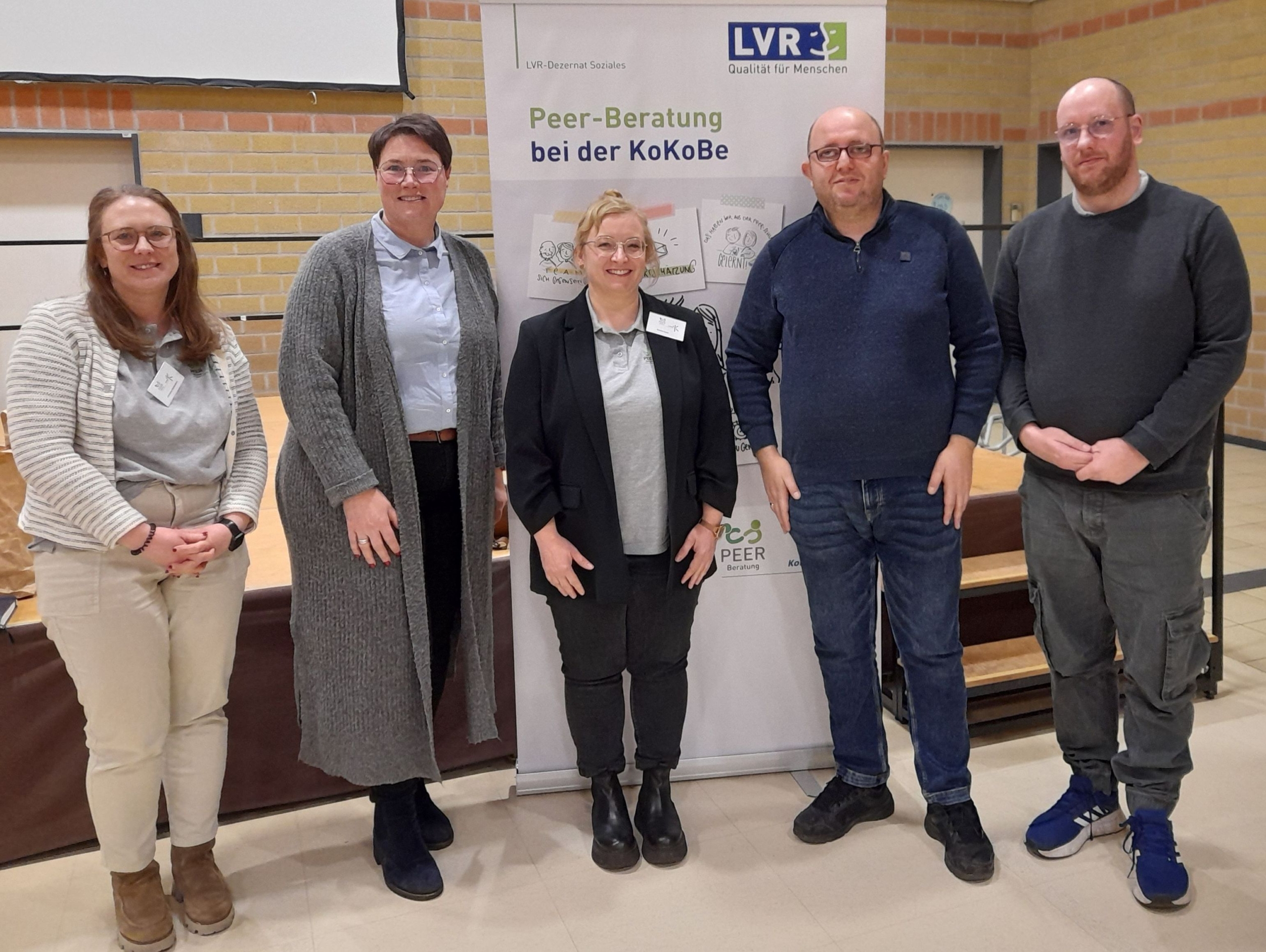 Ein Mann steht vor einer Wand. Er trägt eine Jacke und eine Hose.