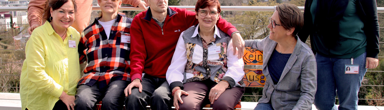 Gruppe von sieben Personen, die fröhlich auf einer Bank im Freien posieren, umgeben von einer landschaftlichen Kulisse. Die Menschen sind unterschiedlich gekleidet und lächeln in die Kamera.