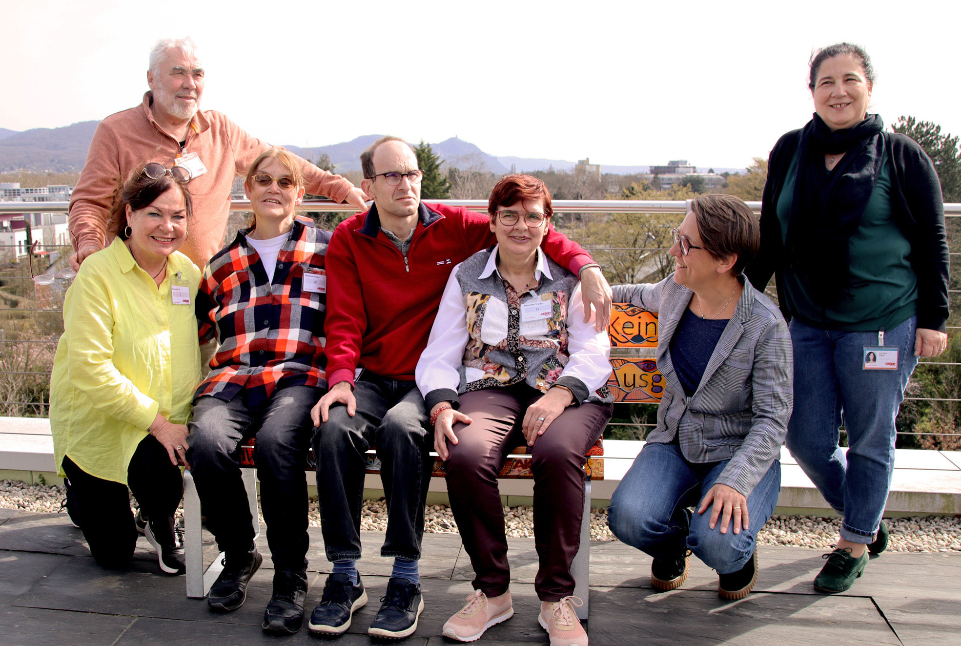 Gruppe von sieben Personen, die fröhlich auf einer Bank im Freien posieren, umgeben von einer landschaftlichen Kulisse. Die Menschen sind unterschiedlich gekleidet und lächeln in die Kamera.