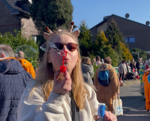 Junge Frau mit Sonnenbrille und blonden Haaren, die auf einem Fest einen roten Luftballon hält. Im Hintergrund sind Menschen und eine sonnige Atmosphäre zu sehen.