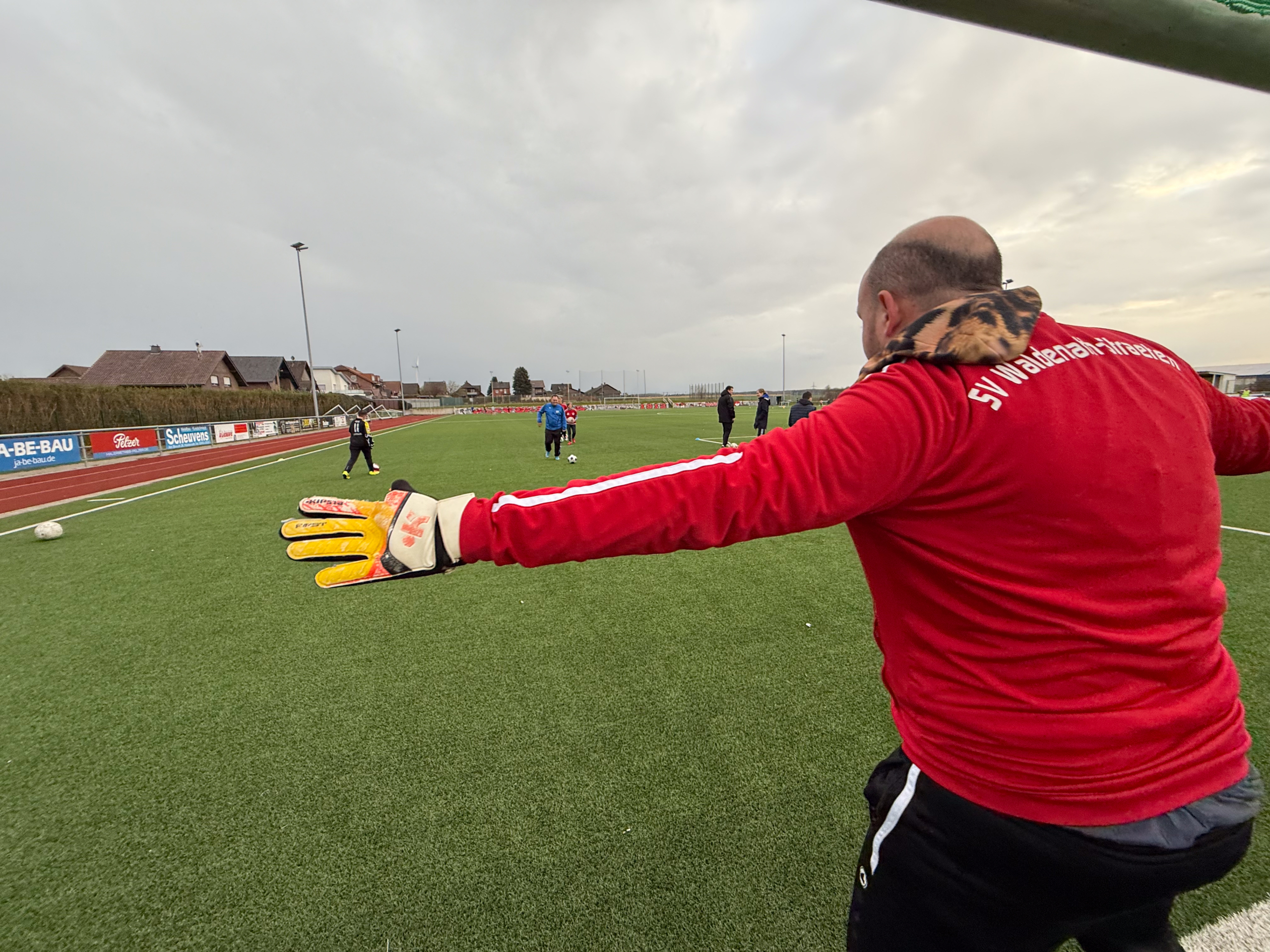 Torwart in rotem Trikot gibt Anweisungen auf dem Fußballplatz während des Trainings. Spieler sind im Hintergrund aktiv.