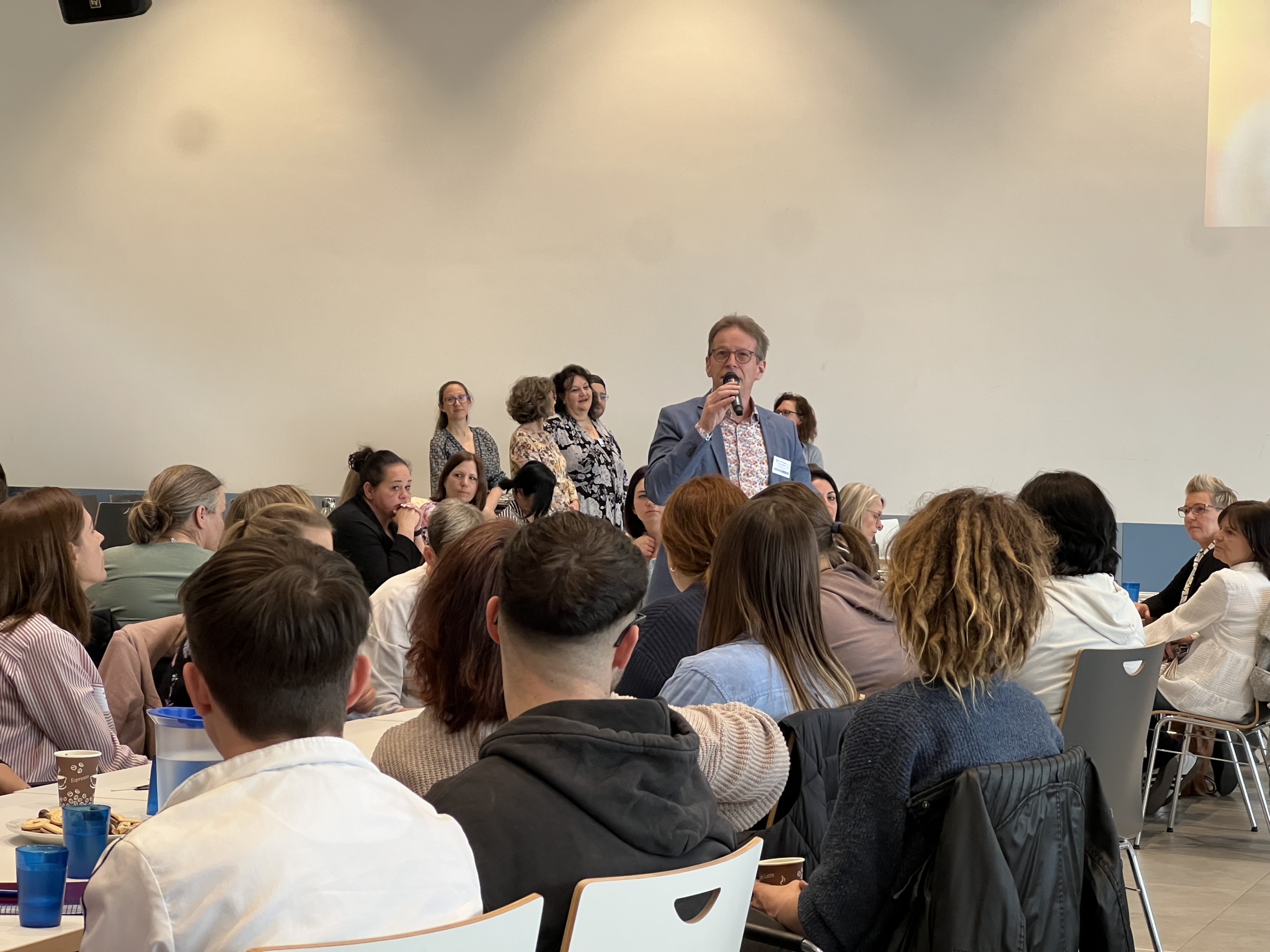 Eine Gruppe von Menschen sitzt in einem Seminarraum und hört einem Redner zu, der eine Präsentation hält. Die Teilnehmer sind aufmerksam und interagieren mit dem Vortrag.