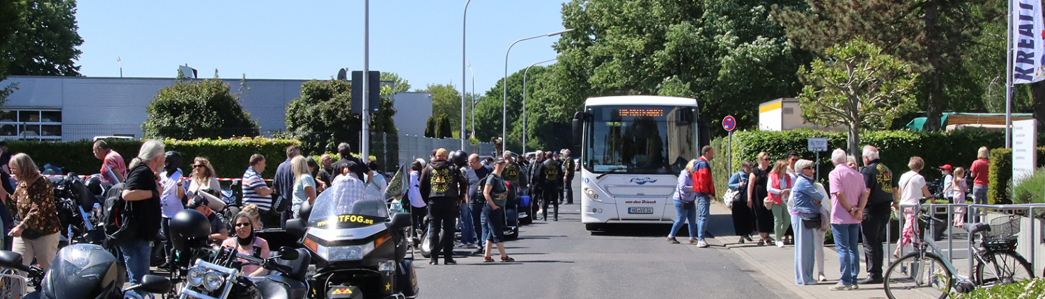 Viele Menschen stehen an einer Straße. Es gibt Motorräder und einen Bus.