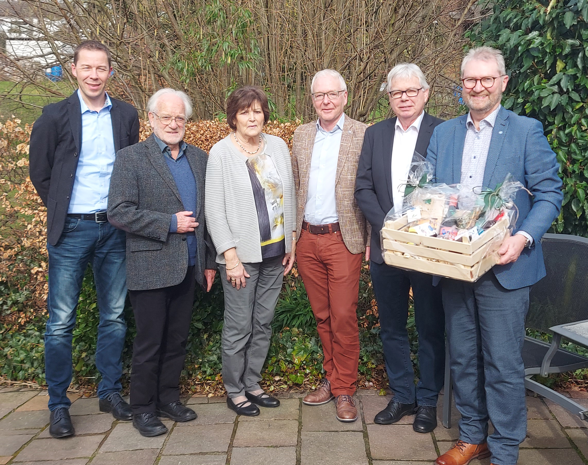 Sechs Personen stehen im Freien vor einem Gebäude mit blättrigem Hintergrund. Vier Männer und zwei Frauen stehen eng nebeneinander, eine Person trägt eine Kiste mit Geschenken.