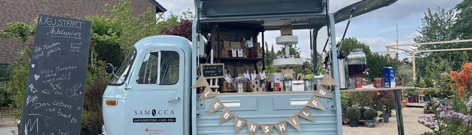 Mobiler Coffee-Shop namens Samocca, der Getränke und Snacks anbietet. Der Wagen ist im Freien aufgestellt, umgeben von Pflanzen und einem Schiefertafel mit freundlichen Botschaften. Ideal für Genuss und Kaffeeerlebnisse in entspannter Atmosphäre.