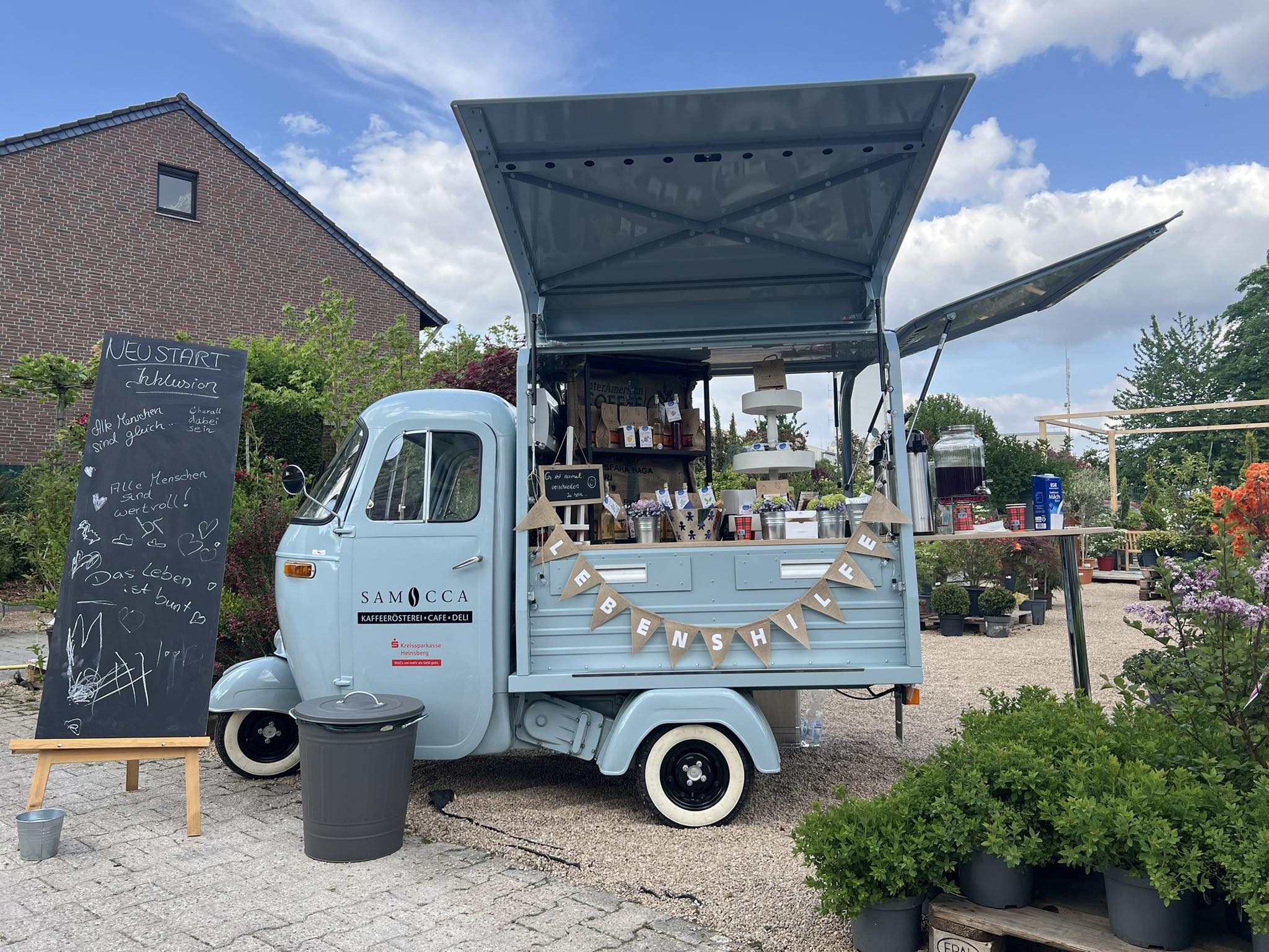 Mobiler Coffee-Shop namens Samocca, der Getränke und Snacks anbietet. Der Wagen ist im Freien aufgestellt, umgeben von Pflanzen und einem Schiefertafel mit freundlichen Botschaften. Ideal für Genuss und Kaffeeerlebnisse in entspannter Atmosphäre.
