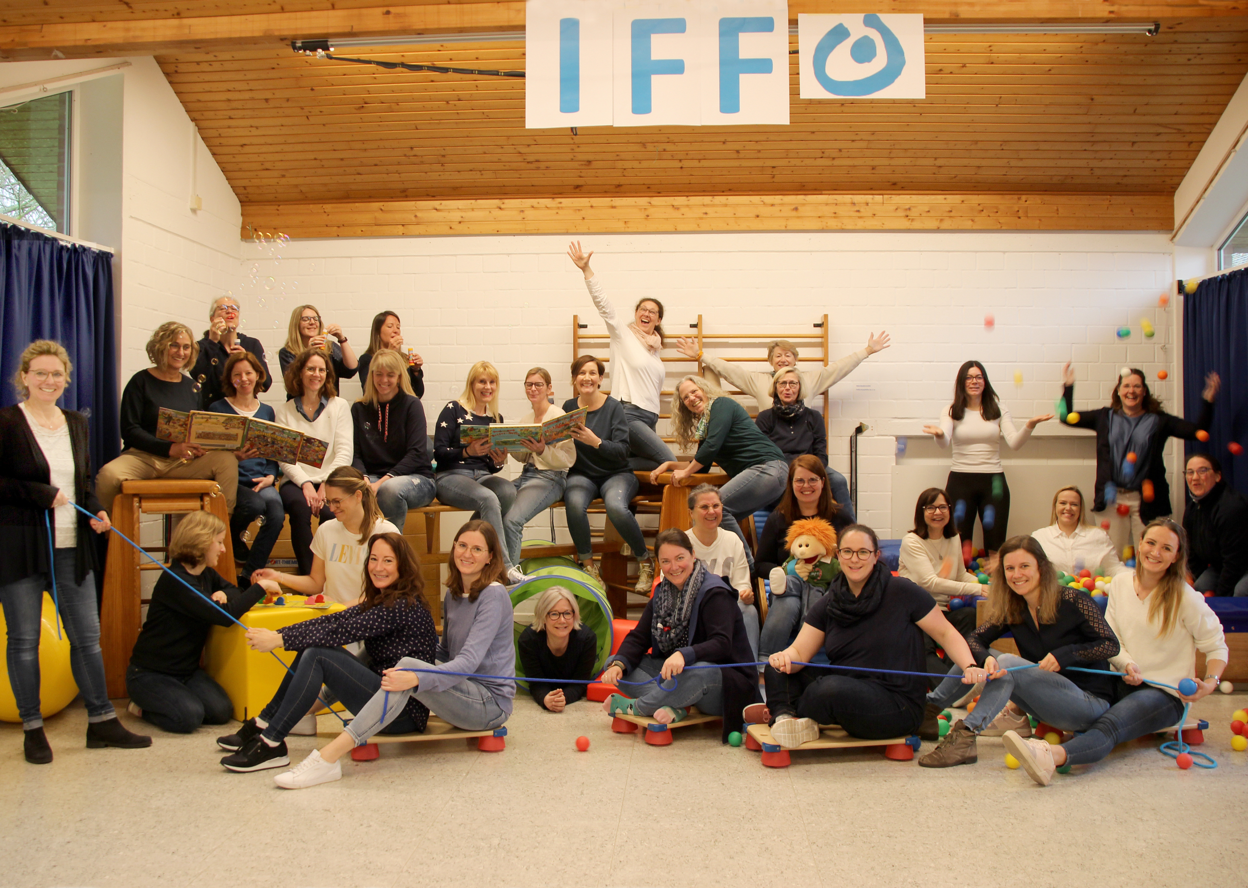 Gruppenbild von Mitarbeitern des IFF, die fröhlich in einem Spielraum posieren. In der Mitte stehen und sitzen Frauen unterschiedlichen Alters, umgeben von Spielzeug und bunten Bällen. Die Atmosphäre ist lebhaft und einladend, was auf eine positive Arbeitskultur hinweist.