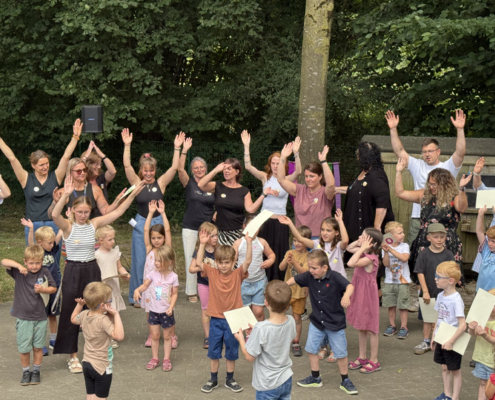 Kinder und Erwachsene tanzen, singen und klatschen gut gelaunt, Sie aben alle die Hände nach oben und singen ein Lied.