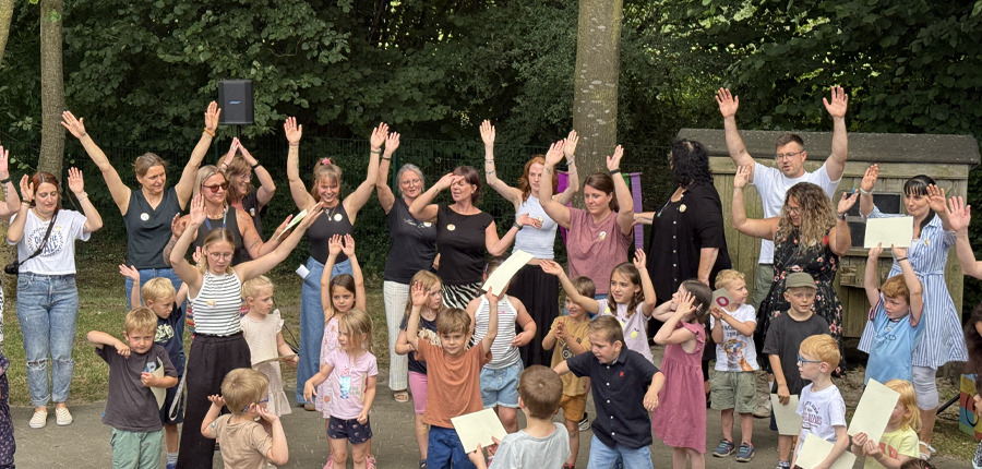 Kinder und Erwachsene tanzen, singen und klatschen gut gelaunt, Sie aben alle die Hände nach oben und singen ein Lied.