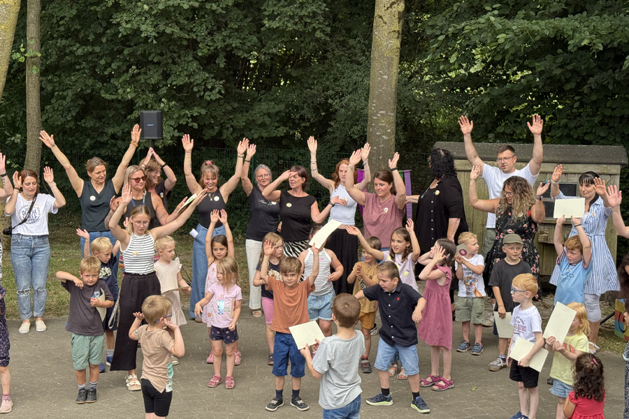Kinder und Erwachsene tanzen, singen und klatschen gut gelaunt, Sie aben alle die Hände nach oben und singen ein Lied.