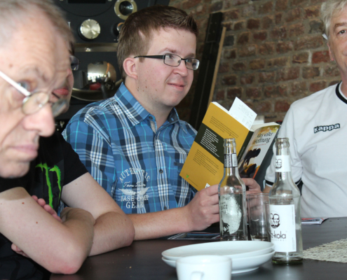 Ein Mann liest ein Buch. Er sitzt an einem Tisch mit Getränken. Im Hintergrund sind zwei weitere Personen zu sehen.