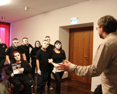 Eine Gruppe von Menschen mit Masken steht in einem Raum. Ein Mann spricht mit ihnen.