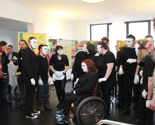 Menschen mit weißen Masken stehen in einem Raum. Einige halten Teller mit Essen. Eine Person sitzt im Rollstuhl.