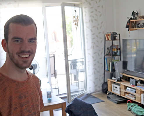 Junger Mann steht im Wohnzimmer und lächelt in die Kamera