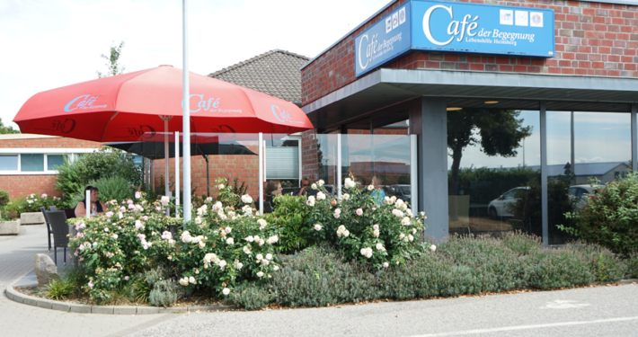 Cafe-der-Begenung-Aussenansicht Aussenaufnahme von einem Cafe. Man sieht Sitzgelegenheiten und große Sonnenschirme. Das Cafe heißt: "Cafe der Begegnung"
