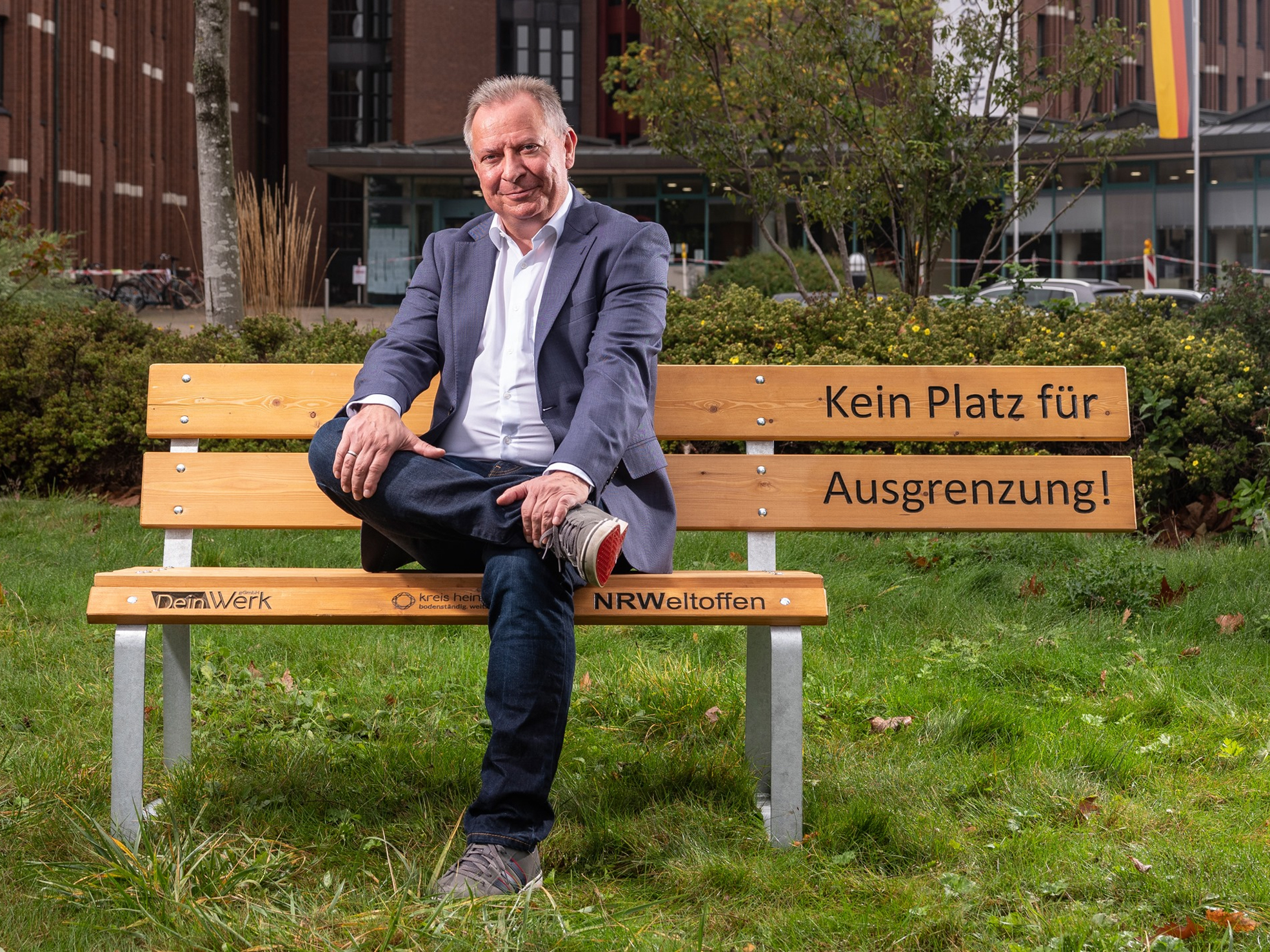Ein Mann sitzt auf einer Bank im Freien. Er trägt einen Anzug und lächelt. Hinter ihm sind Bäume zu sehen.