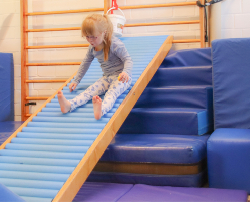 Ein Kind sitzt auf einer blauen, gepolsterten Rutsche in einem Turnraum. Das Kind ist mit Füßen im Socken auf der Rutsche und mit der Hand am Boden festgesetzt. Im Hintergrund ist ein Turngerät aus Holz und eine Sprossleiter zu sehen. Die Umgebung wirkt einladend und für Kinder eingerichtet, wobei der Schwerpunkt auf Bewegung und Aktivität liegt.