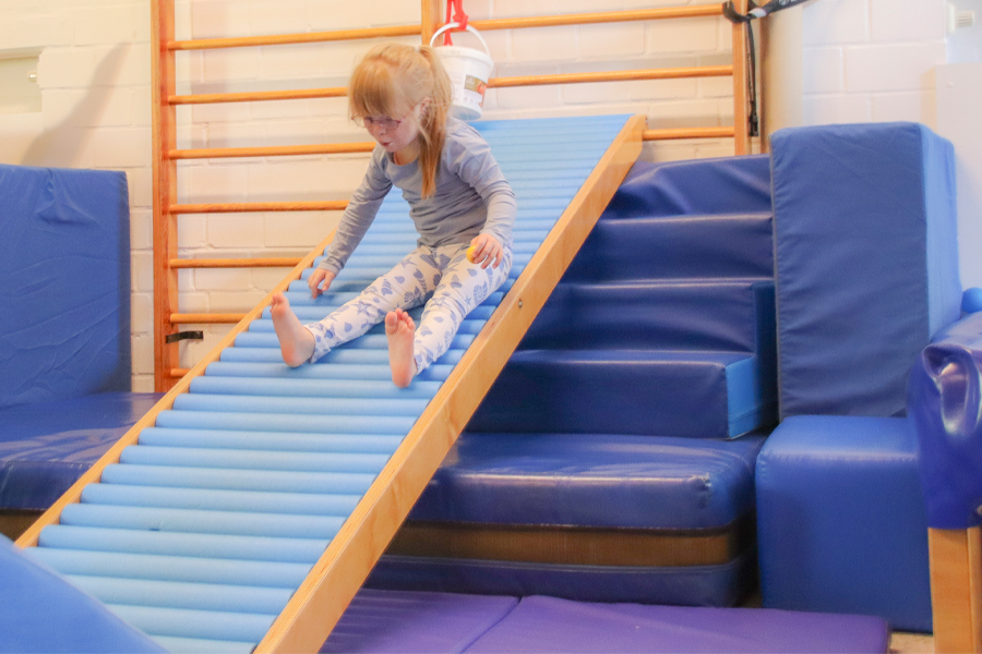 Ein Kind sitzt auf einer blauen, gepolsterten Rutsche in einem Turnraum. Das Kind ist mit Füßen im Socken auf der Rutsche und mit der Hand am Boden festgesetzt. Im Hintergrund ist ein Turngerät aus Holz und eine Sprossleiter zu sehen. Die Umgebung wirkt einladend und für Kinder eingerichtet, wobei der Schwerpunkt auf Bewegung und Aktivität liegt.