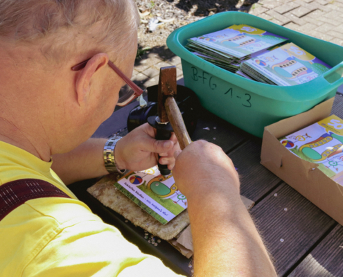 Ein Mann sitzt an einem Tisch und schlägt mit einem Stempel auf Flyer, um sie zu löschern. Auf dem Tisch liegen bereits Flyer mit beschrifteten und verpackten Sets.