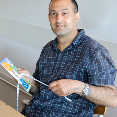 Ein Mann lächelt in die Kamera und hält in seiner hand eine Karte, in der Karte ist ein Schuhriemen eingefädelt, den er mit einer Hand festhält.