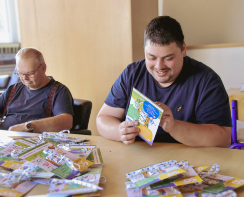 Zwei Männer sitzen an einem Tisch und bereiten Flyer mit Bindfaden vor. Mehrere Flyer sind bereits fertig auf dem Tisch ausgelegt.