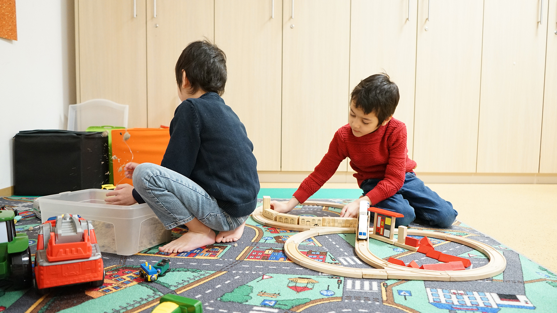 Zwei Kinder spielen gemeinsam auf einem Teppich – eines mit einer Holzeisenbahn, das andere mit einer Kiste voller Sand oder Spielmaterial. Sie wirken konzentriert und vertieft ins Spiel.