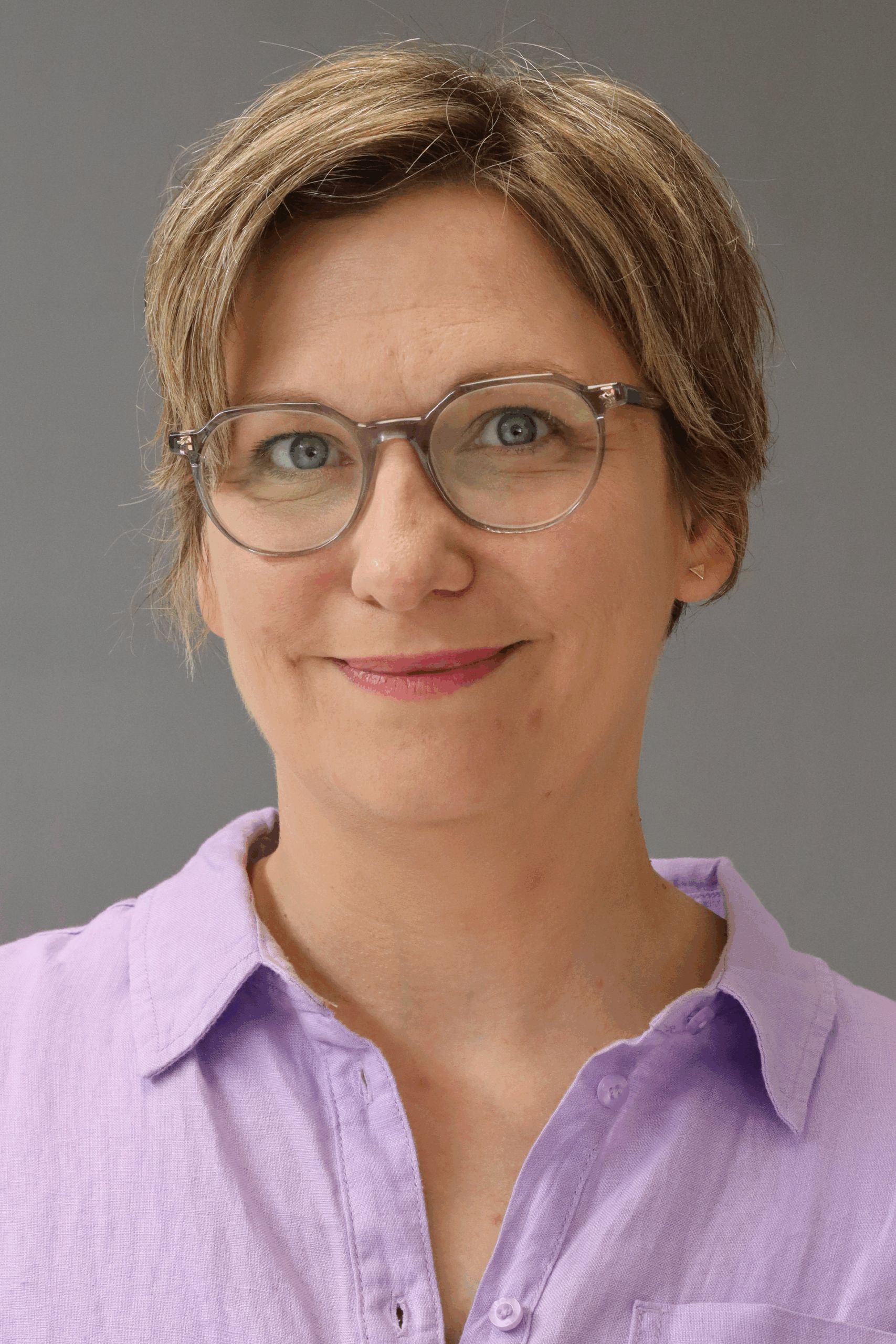 Porträt einer lächelnden Frau mit kurzen, blonden Haaren und Brille, die ein lila Hemd trägt. Der Hintergrund ist grau, was das Gesicht hervorhebt.