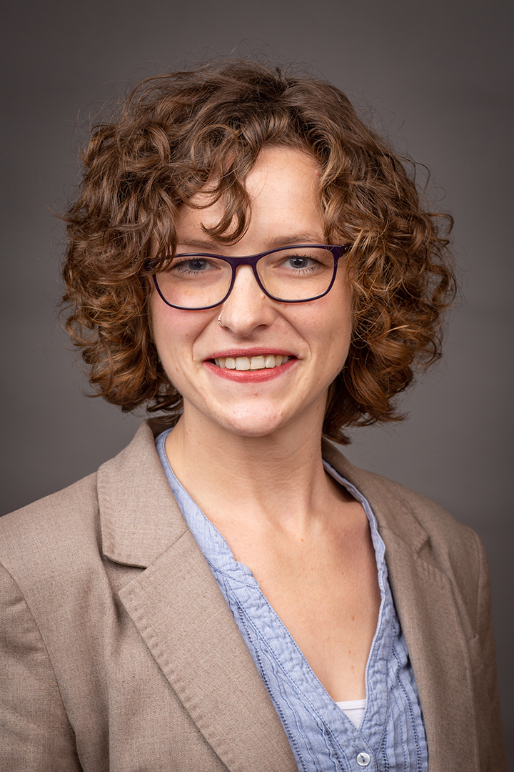 Eine Frau mit Locken und Brille lächelt. Sie trägt eine Jacke und ein blaues Hemd.