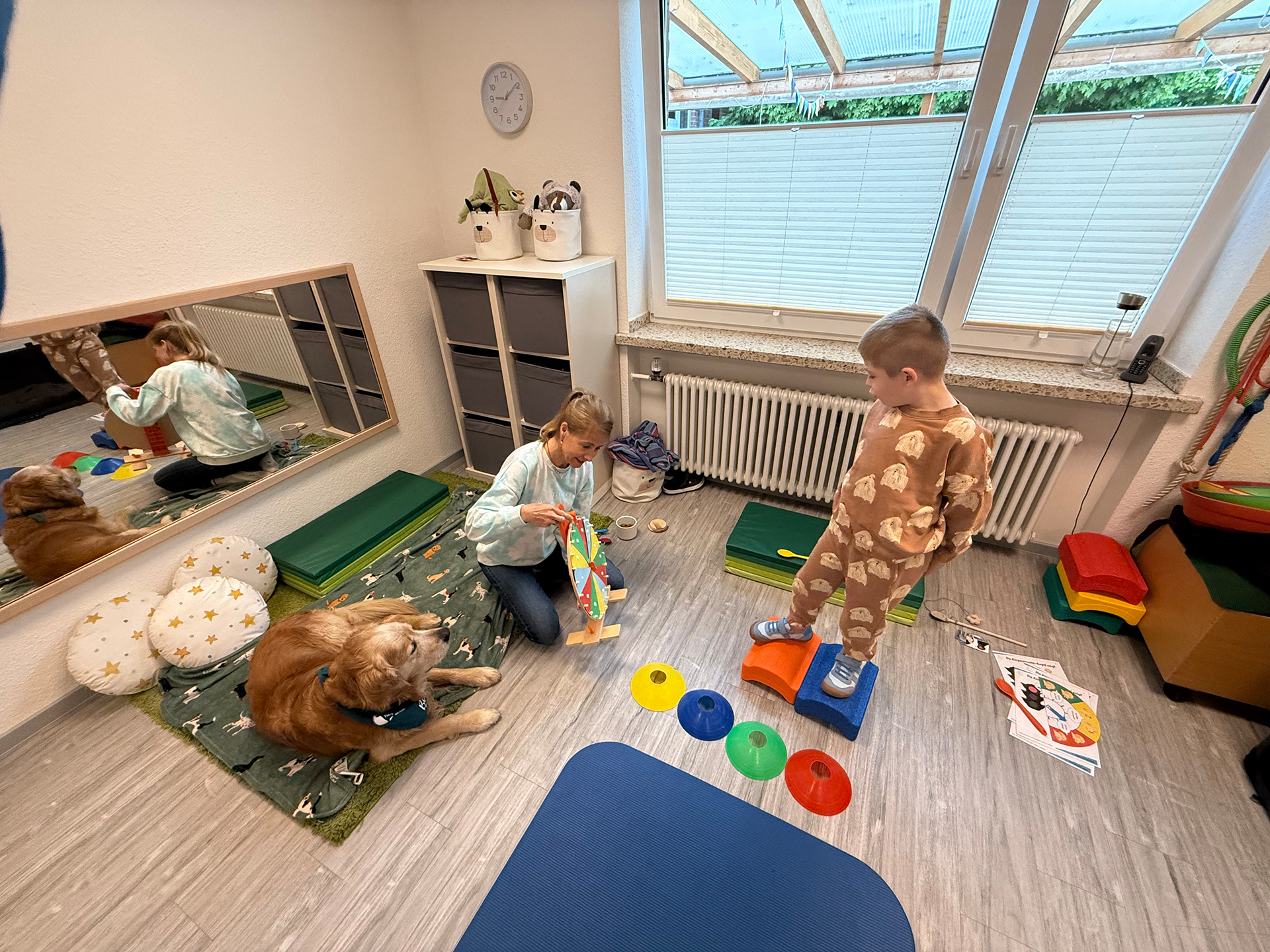 Man sieht in einem Kinder-Therapiezimmer einen Hund, eine Frau und ein Kind. Der kleine Junge steht auf Balanciersteinen und schaut auf eine bunte Drehscheibe, die die Frau in den Händen hälz.