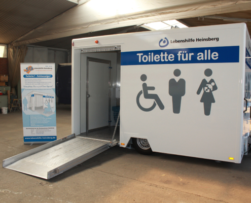 Toilette für Alle (24) Eine mobile Toilette mit einer Rampe. Sie ist weiß und steht auf einem Hof.