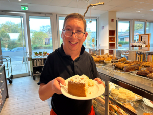 Ein Mann hält einen Kuchen auf einem Teller. Der Kuchen sieht lecker aus. Im Hintergrund sind viele andere Kuchen und Brot zu sehen.