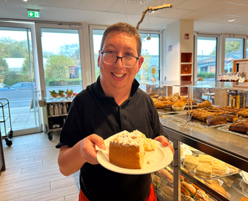 Ein Mann hält einen Kuchen auf einem Teller. Der Kuchen sieht lecker aus. Im Hintergrund sind viele andere Kuchen und Brot zu sehen.