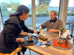 Zwei Personen sitzen an einem Tisch. Eine Person macht Tee. Es gibt Tassen und eine Teekiste.