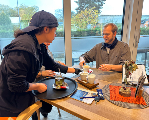 Zwei Personen sitzen an einem Tisch. Eine Person macht Tee. Es gibt Tassen und eine Teekiste.