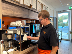 Ein Mann in einer Küche macht Kaffee. Er hat eine Kaffeemaschine bedient und hält eine Tasse.