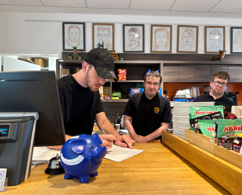 Zwei Männer stehen hinter einem Tresen. Einer schaut auf ein Papier. Es gibt Kaffee und Süßigkeiten.