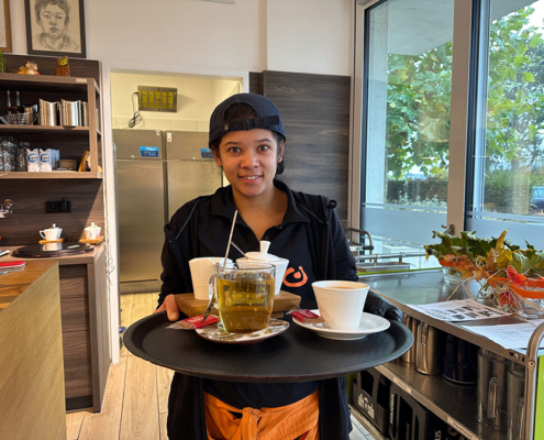 Eine Person hält eine Tablett mit Tassen und einem Tee. Sie steht in einer Küche.
