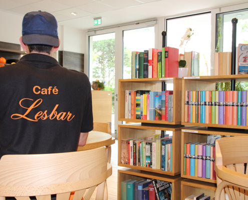 Ein Mann steht in einem Café vor Bücherregalen. Er trägt eine Mütze und ein schwarzes Shirt.