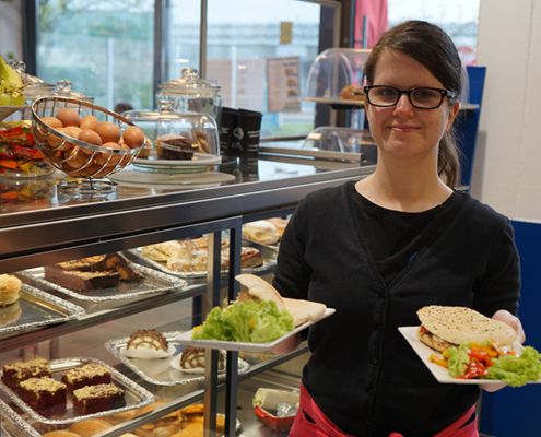 Eine Frau hält Teller mit Sandwiches. Im Hintergrund sind Essen und Kuchen zu sehen.