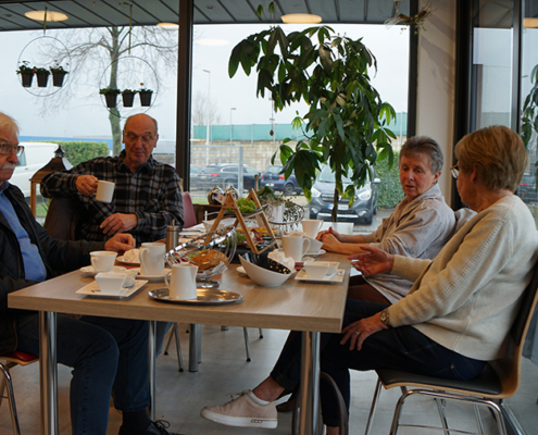 Vier ältere Menschen sitzen an einem Tisch und trinken Kaffee. Sie sitzen in einem hellen Raum mit Blick nach draußen.