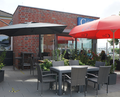 Terrasse mit Tischen und Stühlen. Es gibt Pflanzen und Sonnenschirme. Manche Tische sind leer, andere sind besetzt.