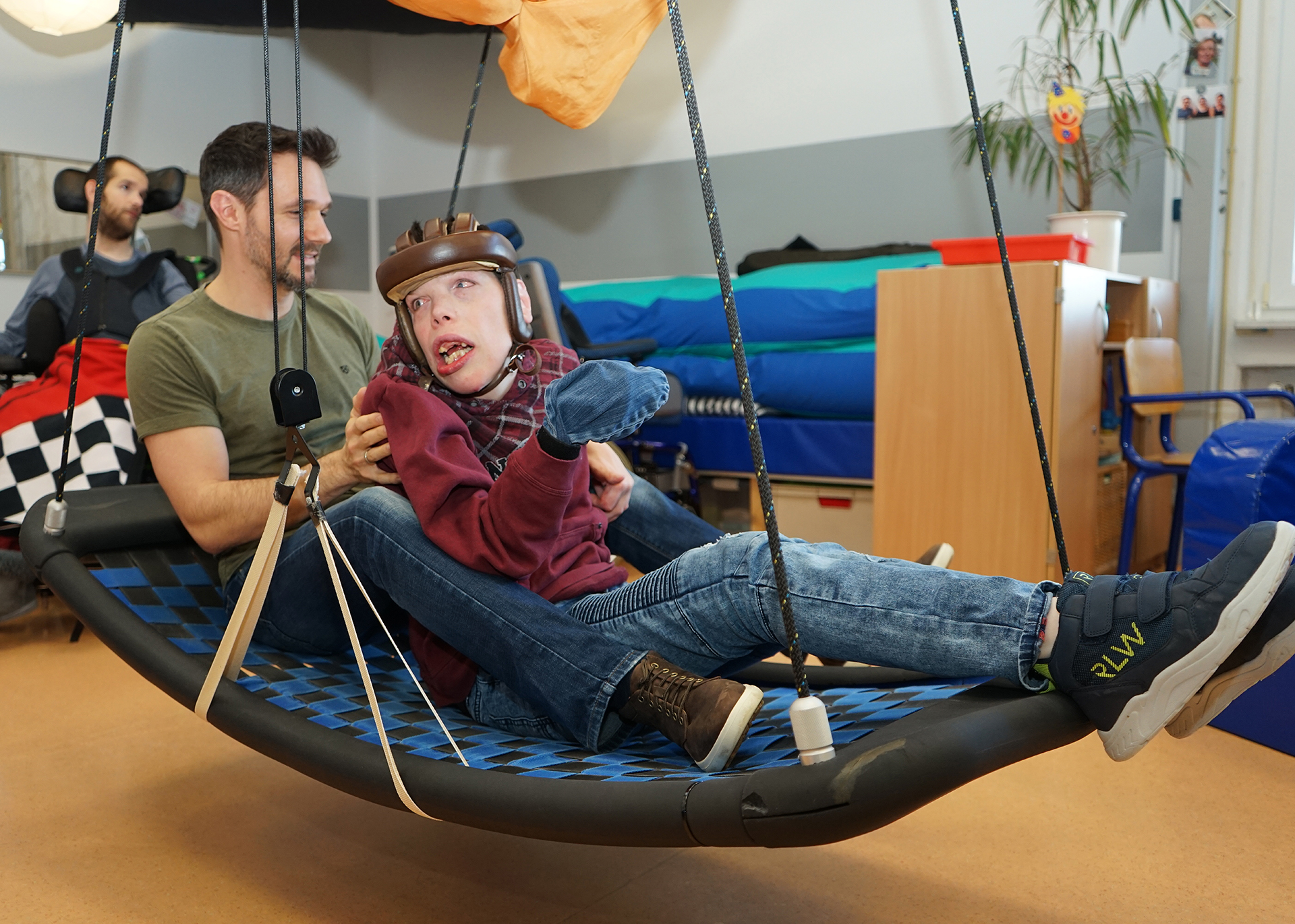 2023_Foerderbereich Ein Kind sitzt in einer Hängematte. Ein Mann steht daneben.