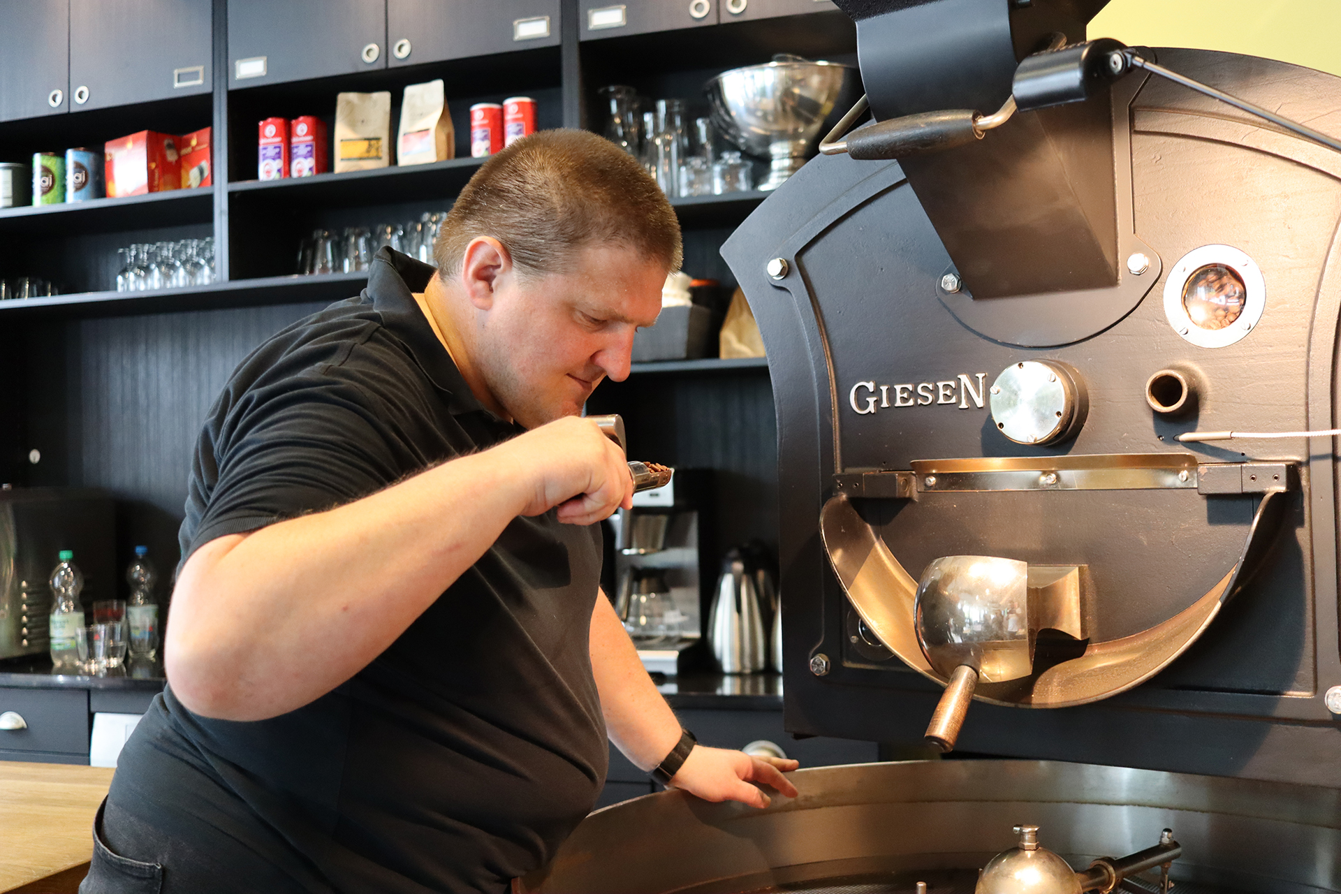 Ein Mann brüht Kaffee. Er schaut in einen Behälter. Im Hintergrund steht eine Kaffeemaschine.